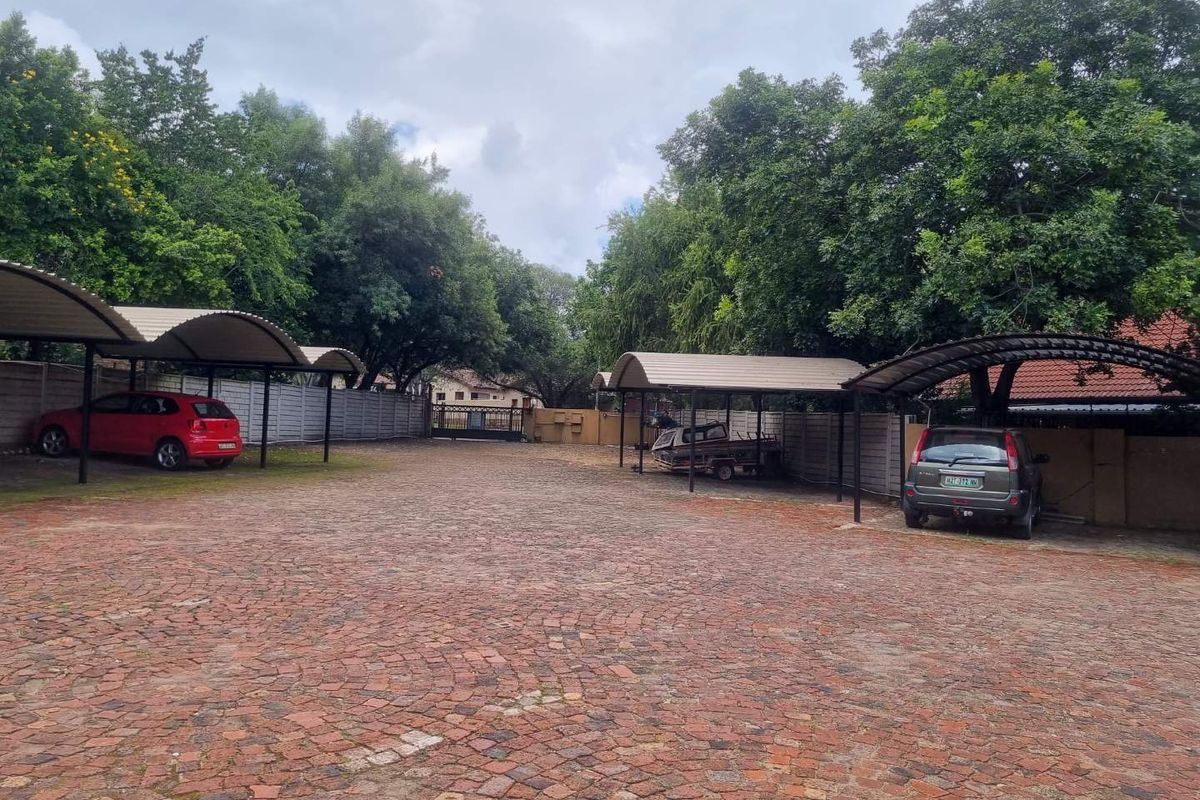 A paved outdoor parking area featuring multiple covered carport bays and perimeter boundary walls, surrounded by mature trees.