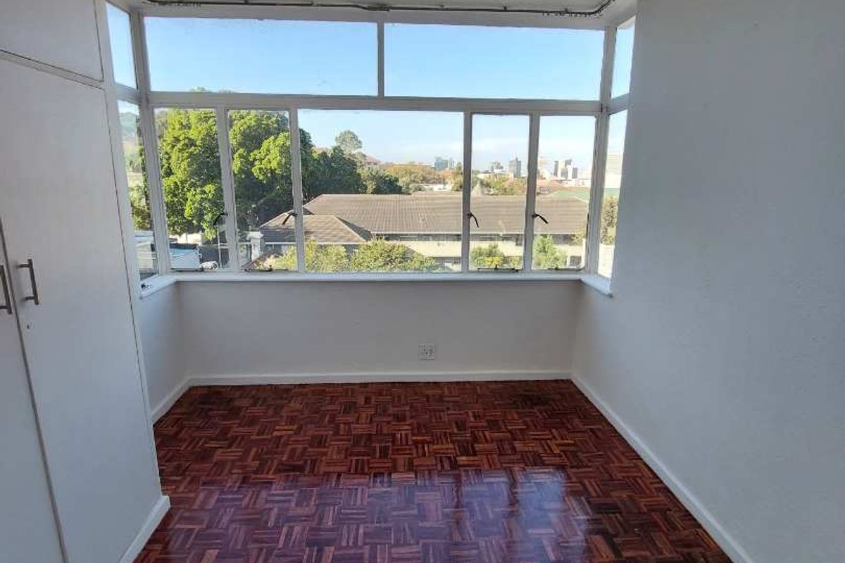 A bright bedroom featuring classic parquet flooring, built-in cupboards, and large windows offering pleasant neighbourhood views.