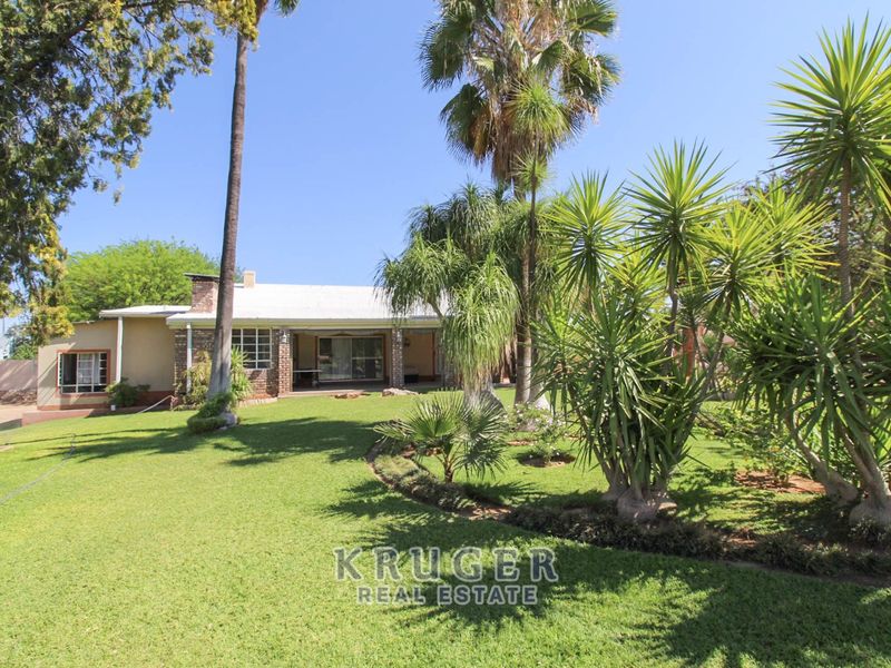 A spacious rear garden featuring a well-maintained lawn, mature palm trees, and a covered patio area attached to the brick-clad residence.