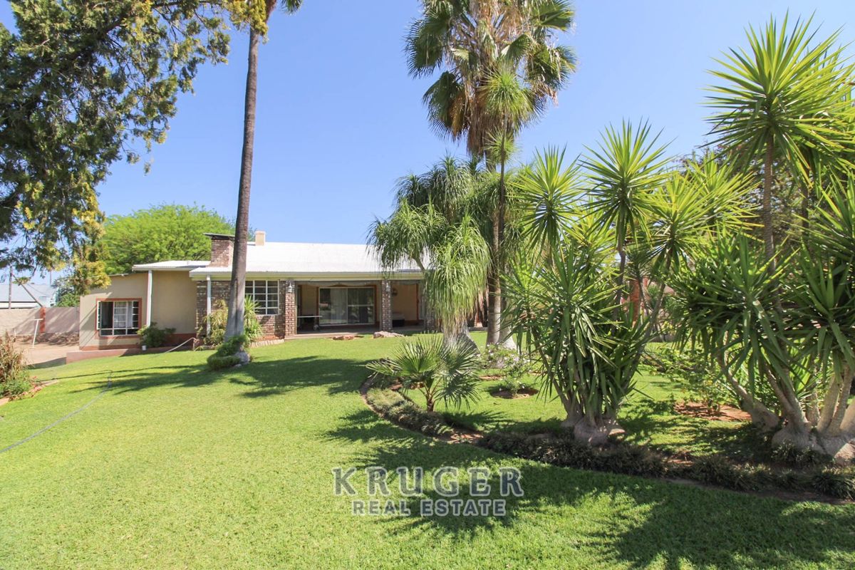 A spacious rear garden featuring a well-maintained lawn, mature palm trees, and a covered patio area attached to the brick-clad residence.