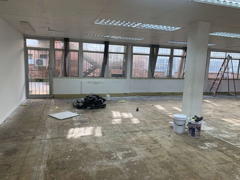 Open-plan industrial office space featuring large windows, suspended ceilings, and a structural column, currently undergoing renovation with exposed flooring.