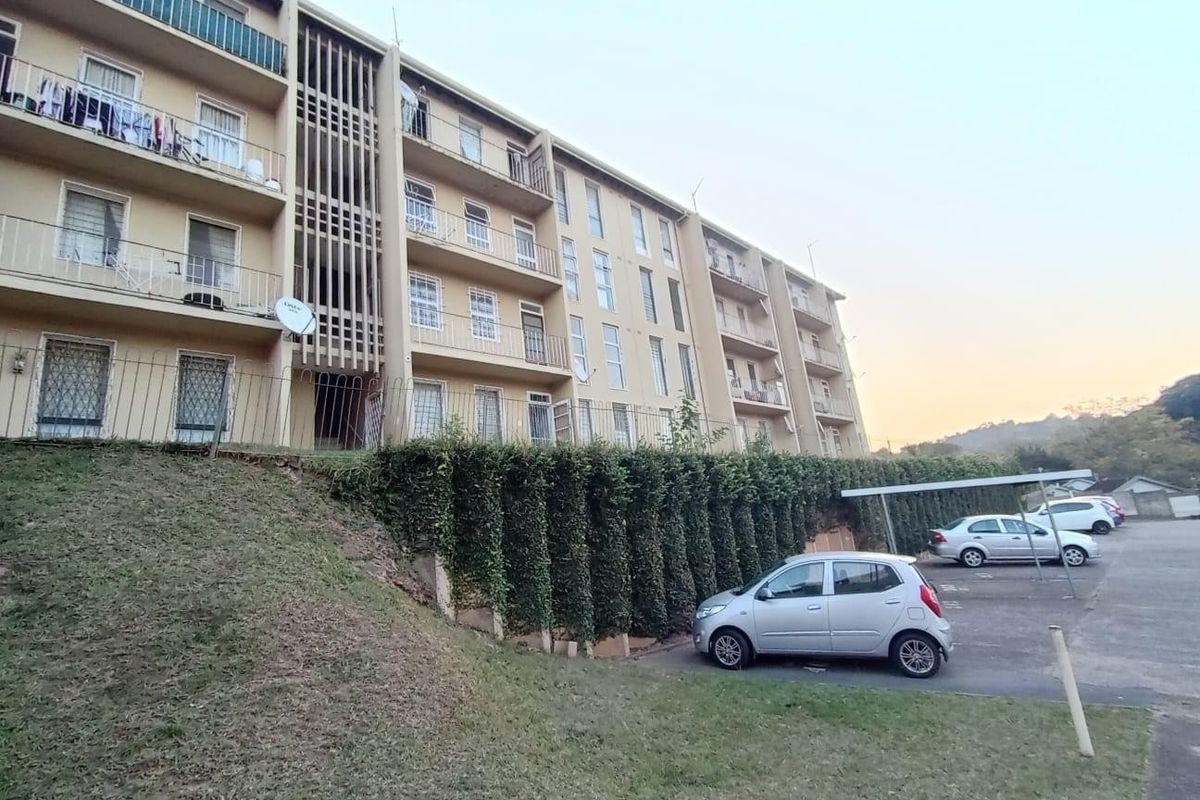 Exterior view of a multi-story apartment building featuring private balconies, security gates, and a paved parking area with carports.