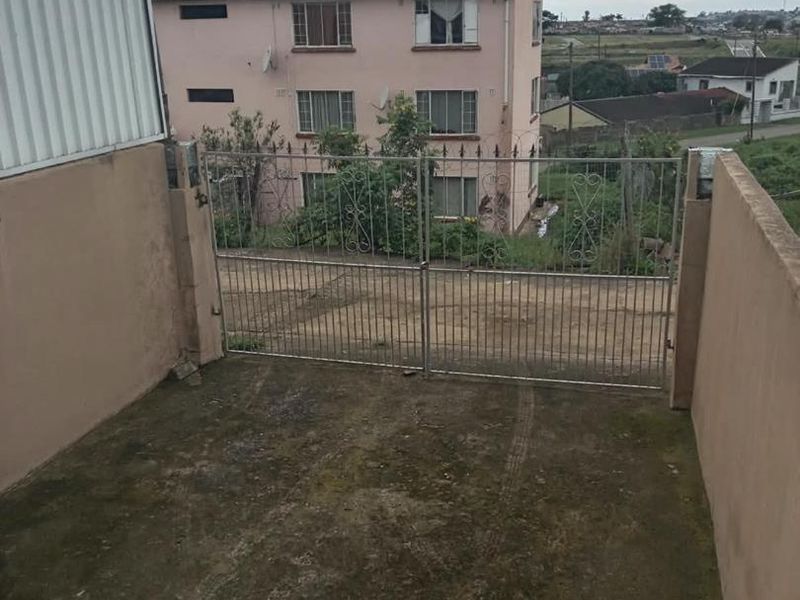 An exterior view of a concrete driveway entrance secured by a metal gate, showing surrounding residential property and boundary walls.