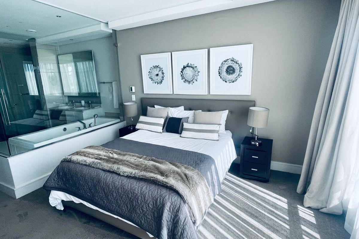 A modern bedroom featuring an integrated glass-enclosed en-suite bathroom with a soaking tub, neutral wall tones, and recessed ceiling lighting.