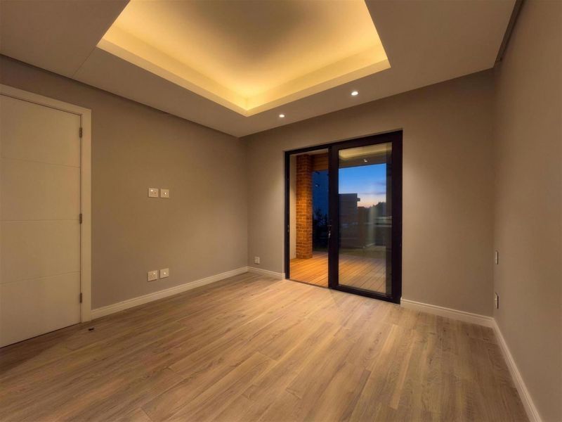 A modern, empty bedroom featuring a recessed ceiling with cove lighting, laminate flooring, and a sliding glass door leading to an outdoor area.