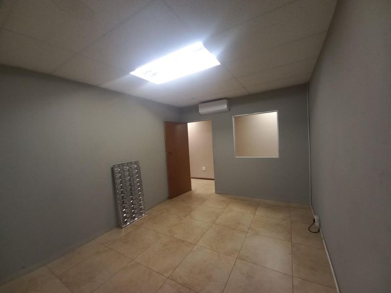 A vacant office space featuring tiled flooring, a suspended ceiling with recessed lighting, a wall-mounted air conditioning unit, and an internal viewing window.