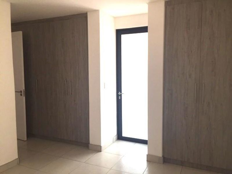 A bright, empty bedroom featuring modern wood-finish built-in cupboards, neutral tiled flooring, and a glass-panelled door leading to an outdoor area.