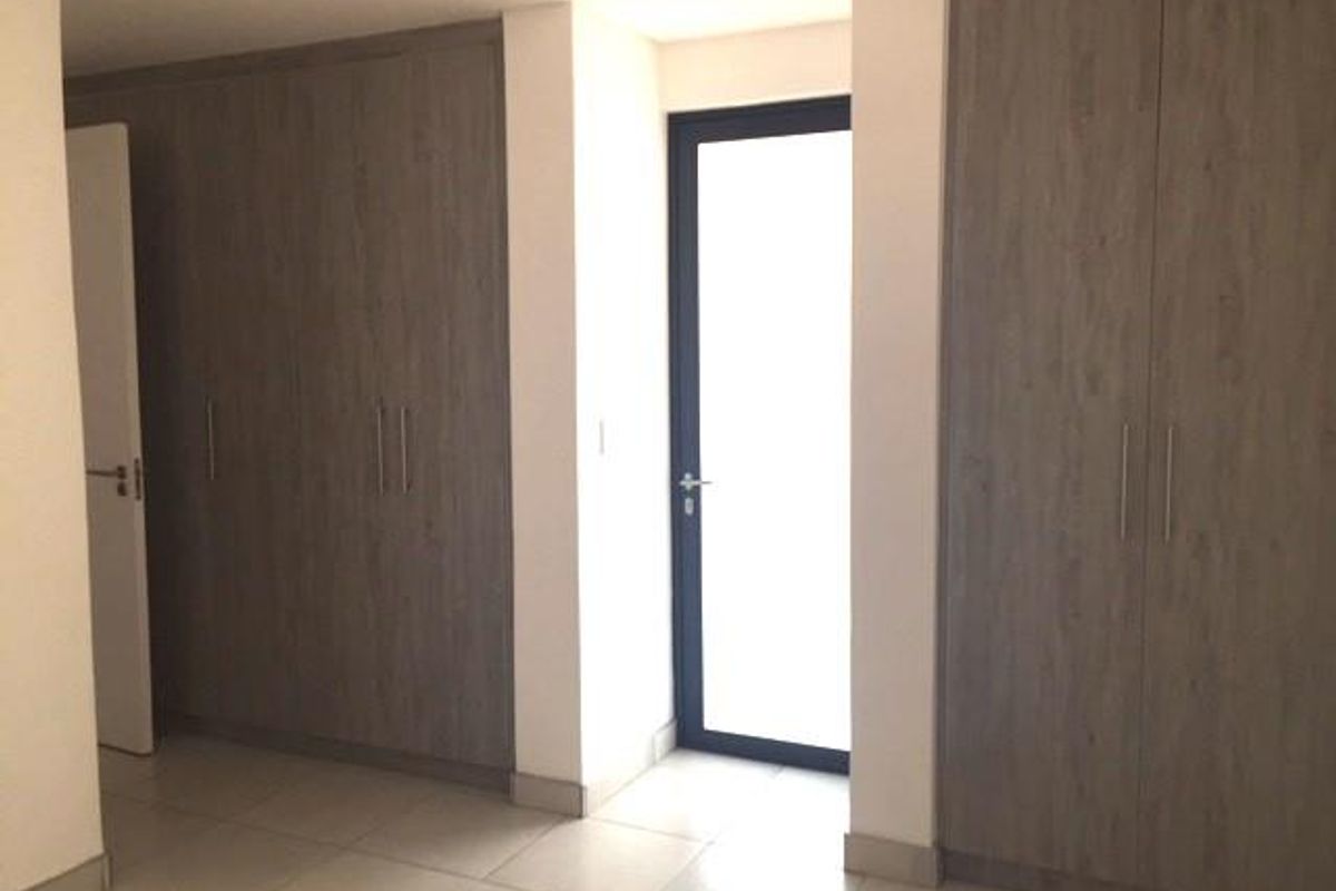 A bright, empty bedroom featuring modern wood-finish built-in cupboards, neutral tiled flooring, and a glass-panelled door leading to an outdoor area.
