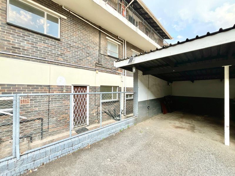 A covered carport area adjacent to a brick apartment building, featuring a security gate and fenced perimeter.