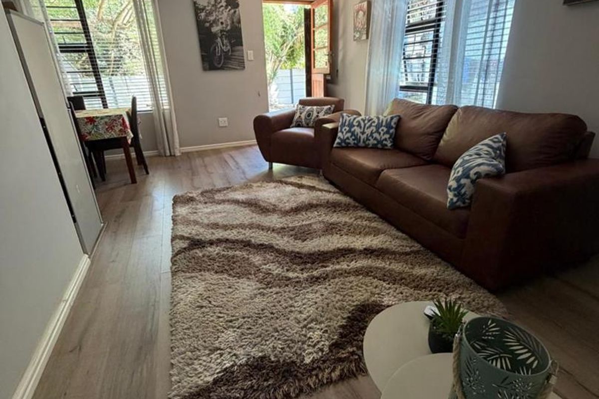 A bright living room featuring wood-look laminate flooring, large windows with blinds, and direct access to the outdoor area.