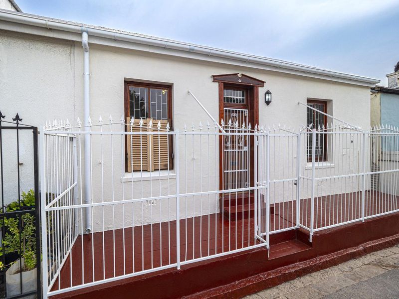 Front facade of a cottage featuring a secure gated entrance, timber-framed windows with security bars, and a painted concrete porch area.