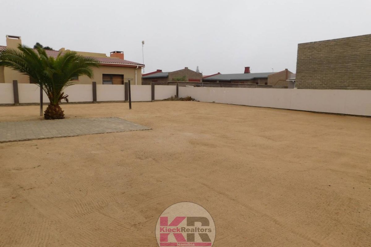 A spacious, low-maintenance rear garden area featuring a sandy surface, perimeter boundary walls, and a small paved section.