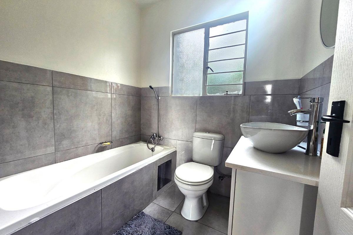 A modern bathroom featuring grey wall and floor tiles, a built-in bathtub with shower attachment, a toilet, and a vessel sink on a vanity.