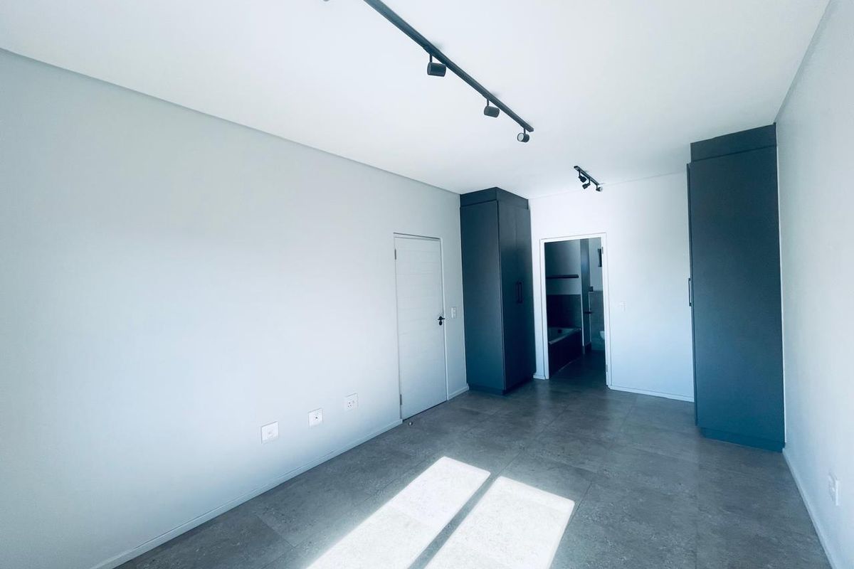 A modern, empty bedroom featuring sleek dark built-in cupboards, contemporary track lighting, and large-format floor tiles, with an adjoining bathroom visible.