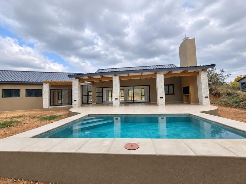 A modern outdoor entertainment area featuring a swimming pool, covered patio with exposed timber beams, stone-clad pillars, and a built-in fireplace.