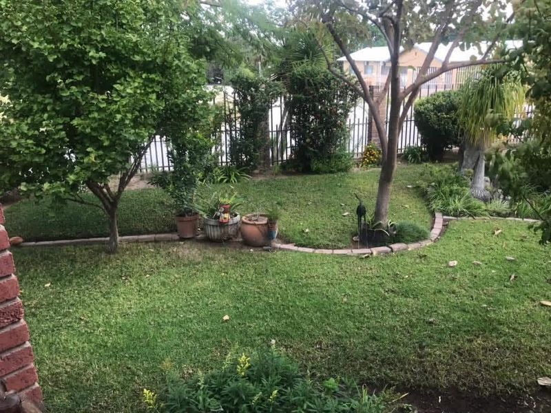 A private garden area featuring a well-maintained lawn, mature trees, and brick-edged garden beds enclosed by a metal perimeter fence.