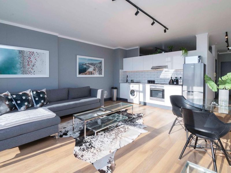 Modern open-plan living area featuring laminate flooring, track lighting, and a contemporary kitchen with subway tile backsplash and integrated appliances.