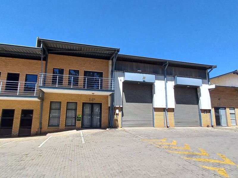 Front elevation of an industrial complex featuring face brick construction, roller shutter doors, and upper-level balcony access with paved parking area.