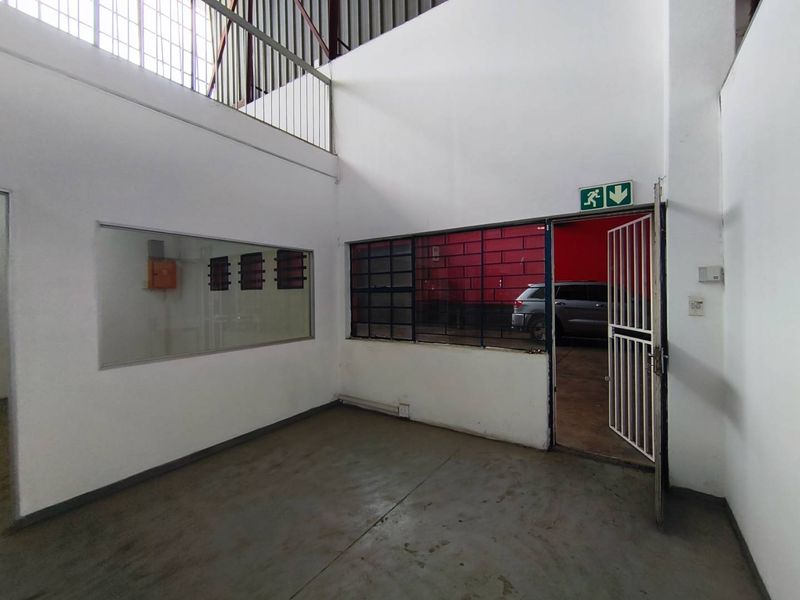 An empty industrial office space featuring concrete flooring, steel-framed windows, and a mezzanine level with a security gate entrance.