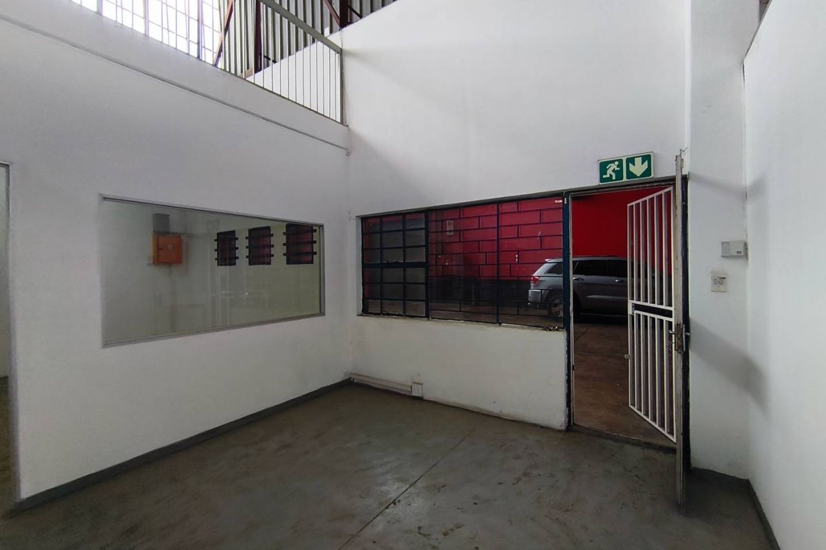 An empty industrial office space featuring concrete flooring, steel-framed windows, and a mezzanine level with a security gate entrance.