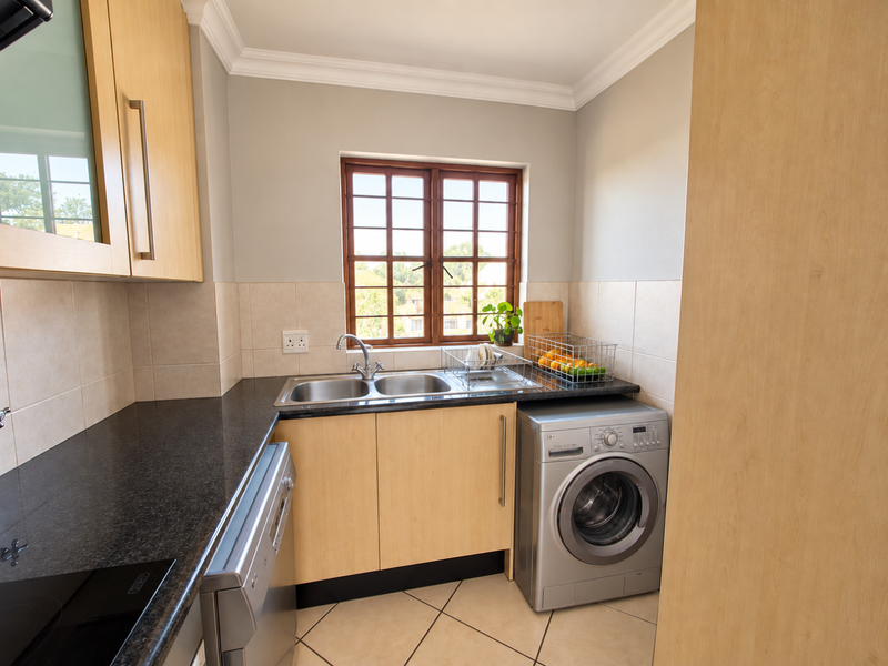 A functional scullery featuring light wood cabinetry, dark granite countertops, a double sink, and tiled finishes, providing efficient space for laundry and kitchen prep.