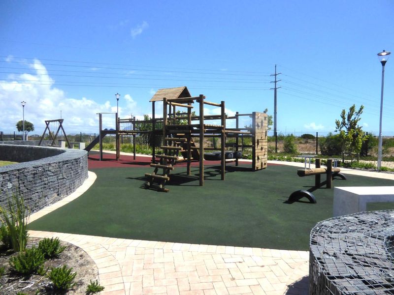 A well-maintained outdoor playground area featuring modern timber play structures, rubberized safety flooring, and decorative gabion stone retaining walls.