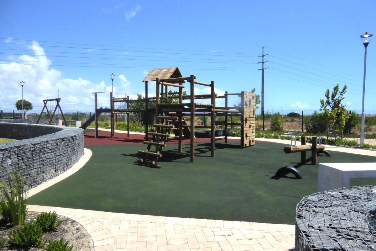 A well-maintained outdoor playground area featuring modern timber play structures, rubberized safety flooring, and decorative gabion stone retaining walls.