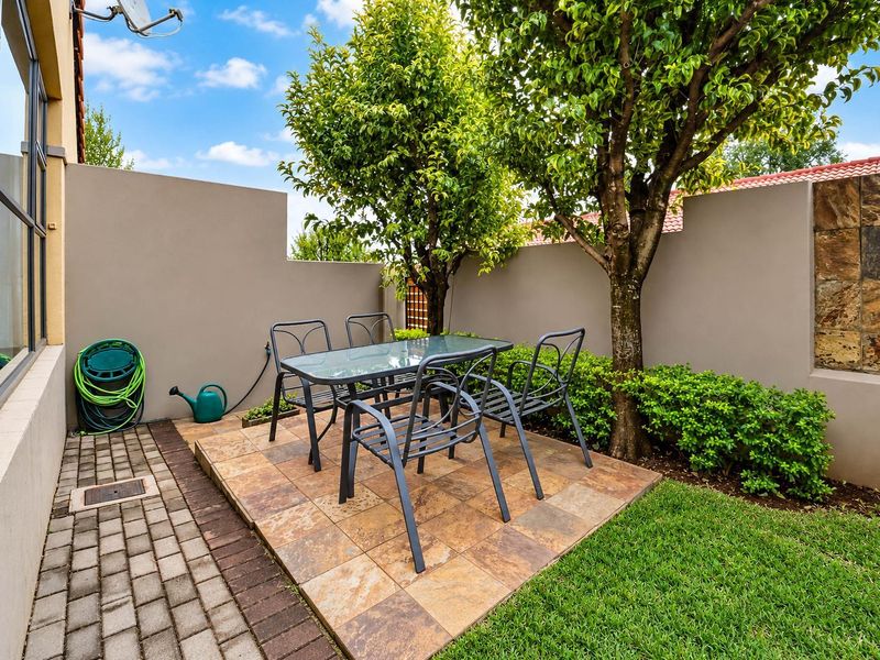A private rear garden featuring a paved patio area, established trees, and a well-maintained lawn enclosed by a solid boundary wall.