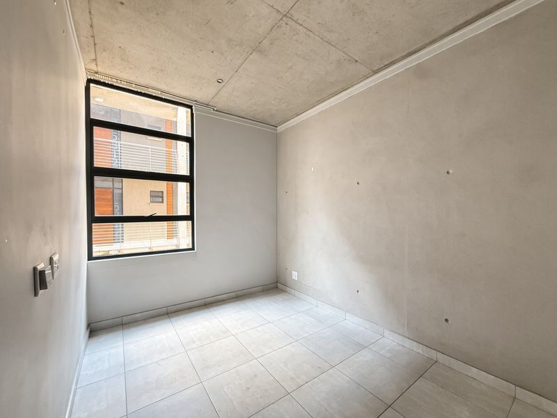 A bright, empty bedroom featuring an industrial-style exposed concrete ceiling, neutral tiled flooring, and a large window providing natural light.