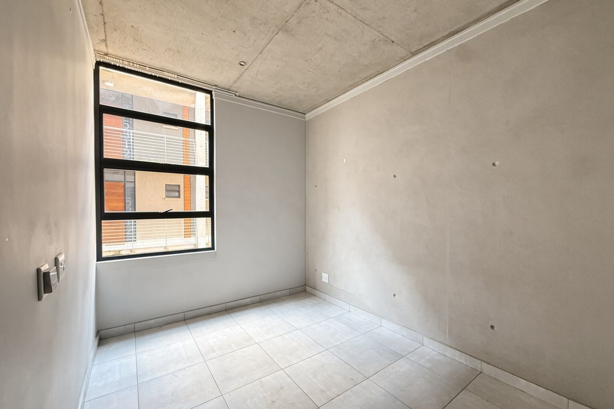A bright, empty bedroom featuring an industrial-style exposed concrete ceiling, neutral tiled flooring, and a large window providing natural light.
