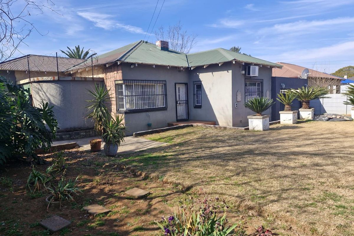 Rear view of a single-story house featuring a corrugated metal roof, security bars on windows, and a spacious garden area.
