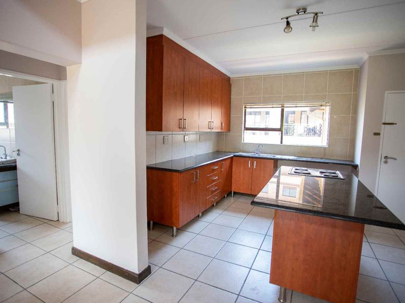 A spacious, empty kitchen featuring wooden cabinetry, granite countertops, a central island with a built-in hob, and neutral ceramic floor tiles.