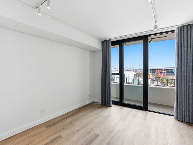 A bright, empty bedroom featuring modern laminate flooring, track lighting, and a large sliding glass door providing access to a private balcony.
