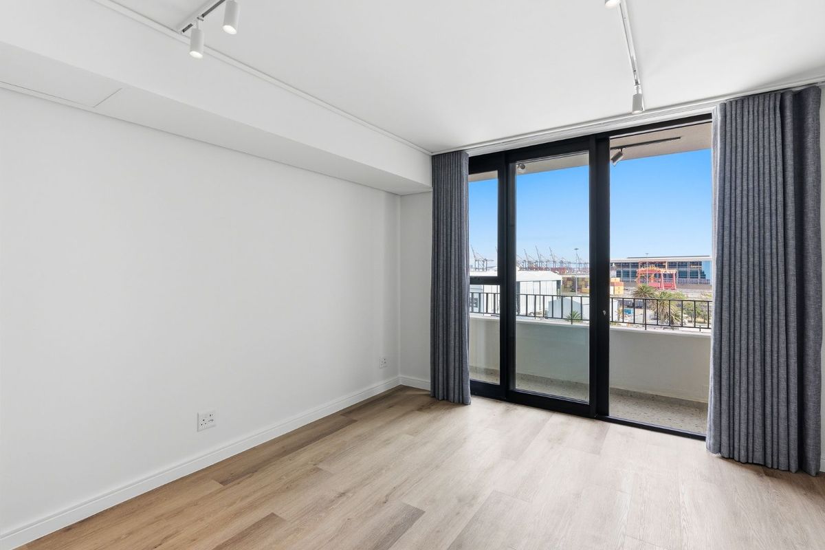 A bright, empty bedroom featuring modern laminate flooring, track lighting, and a large sliding glass door providing access to a private balcony.
