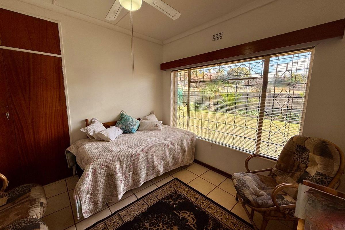A bedroom featuring tiled flooring, built-in wooden cupboards, a ceiling fan, and large windows with security bars overlooking the garden.