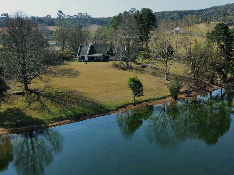 Aerial view of a farm property featuring a dwelling situated on a large lawn adjacent to a scenic dam and surrounding rural landscape.