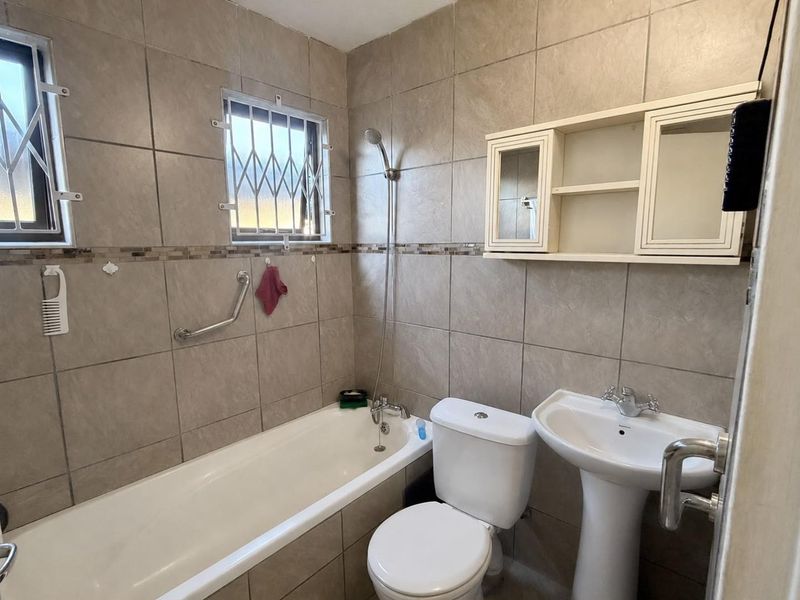 A functional bathroom featuring a bathtub, pedestal basin, toilet, and wall-mounted storage cabinet, finished with neutral wall tiling and security-fitted windows.