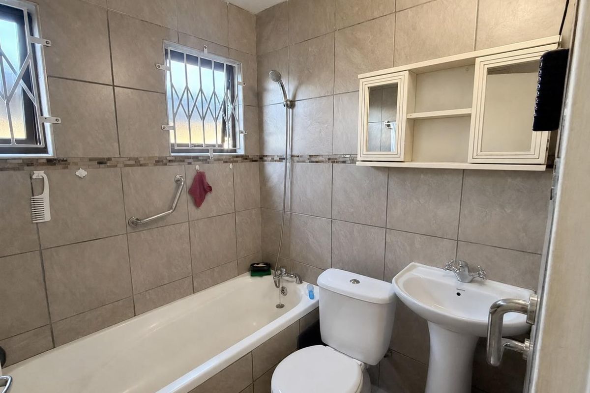 A functional bathroom featuring a bathtub, pedestal basin, toilet, and wall-mounted storage cabinet, finished with neutral wall tiling and security-fitted windows.