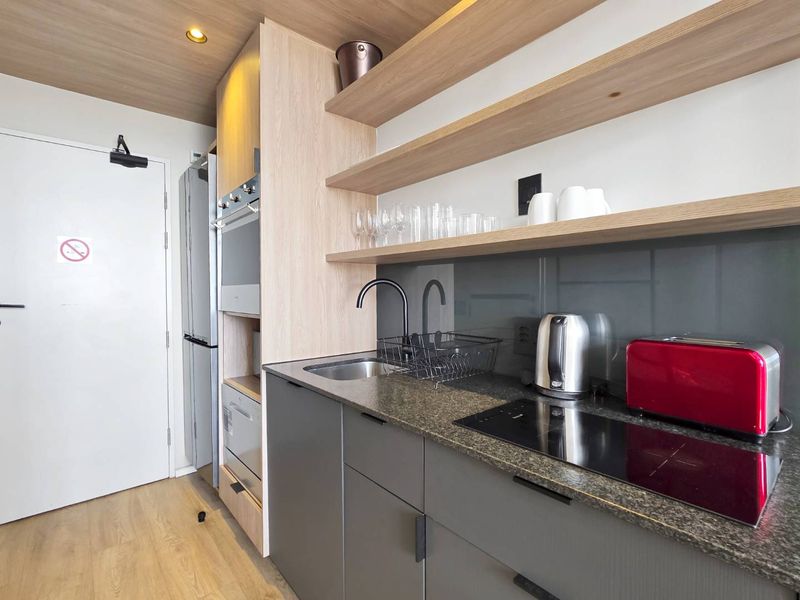 Modern compact kitchen featuring sleek grey cabinetry, stone countertops, built-in oven, induction hob, and stylish floating timber shelving.
