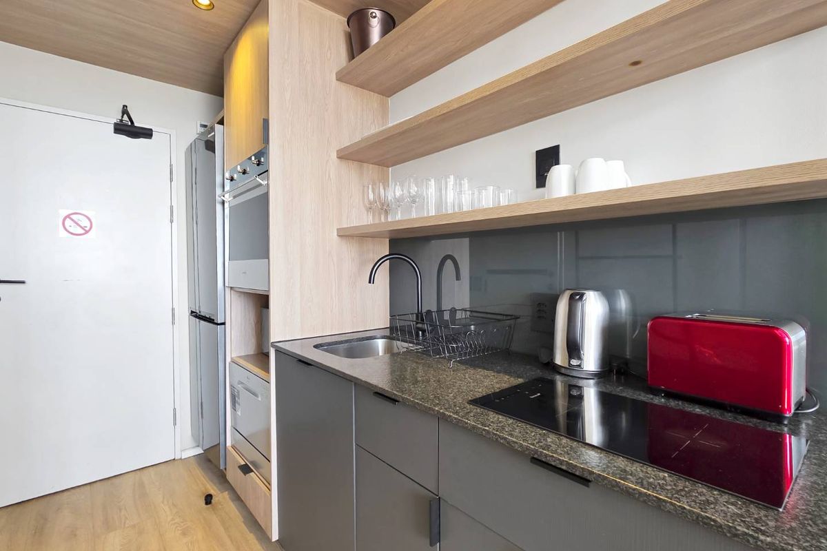 Modern compact kitchen featuring sleek grey cabinetry, stone countertops, built-in oven, induction hob, and stylish floating timber shelving.