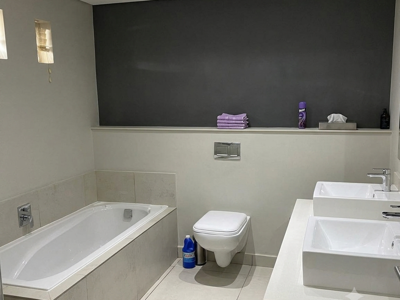 Modern bathroom featuring a built-in bathtub, wall-hung toilet, and a dual vanity unit with contemporary basin fixtures and neutral tiling.