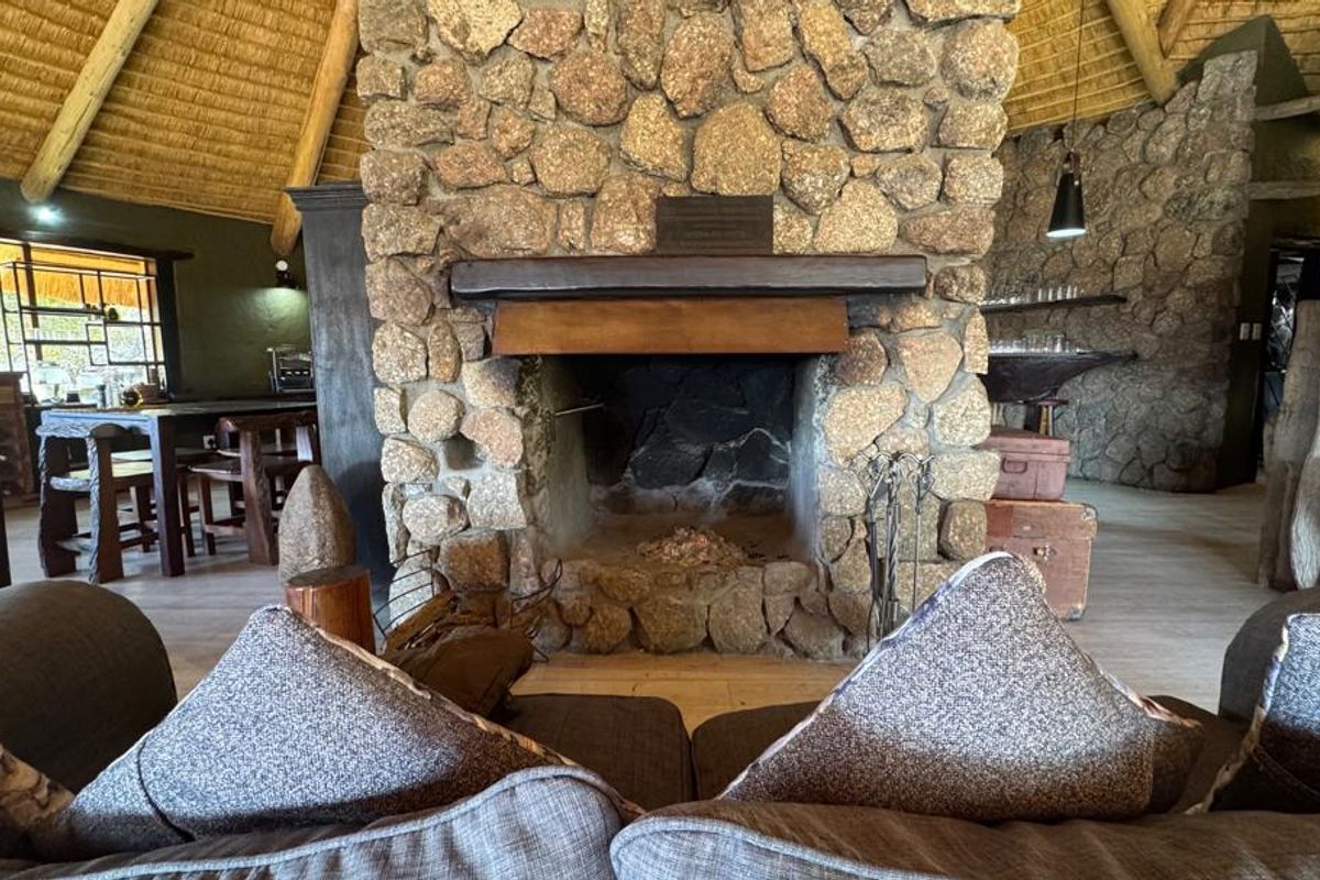 Spacious lodge living area featuring a prominent central stone fireplace, high thatched ceilings with exposed timber beams, and rustic stone wall finishes.