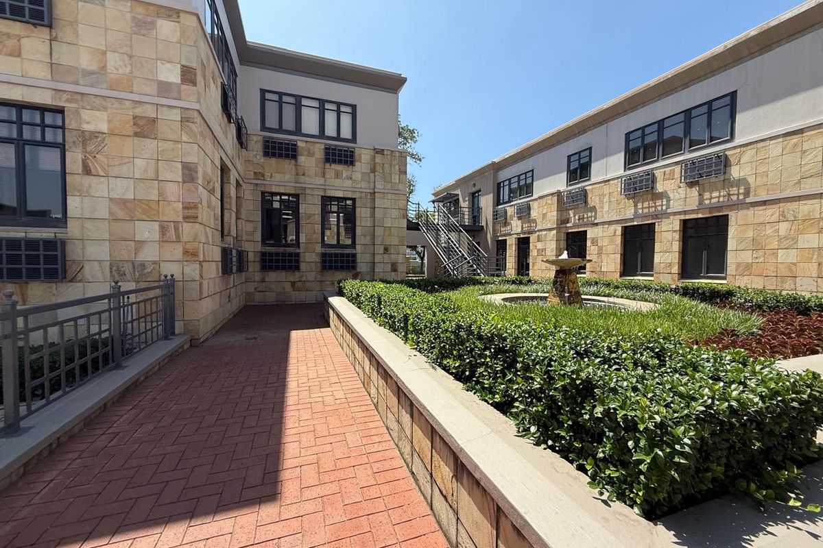 A well-maintained office courtyard featuring stone-clad building facades, a paved walkway, landscaped garden beds, and a central water fountain.