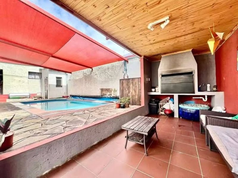 Outdoor entertainment area featuring a swimming pool, built-in braai, tiled patio, and timber ceiling with a red shade sail.