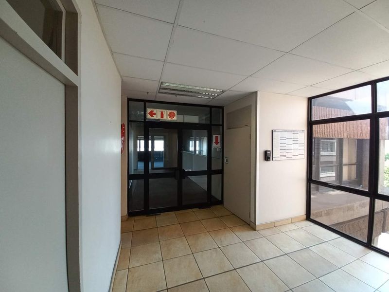 A bright office building hallway featuring tiled flooring, a suspended ceiling with recessed lighting, and large glass partitions leading to adjacent office spaces.