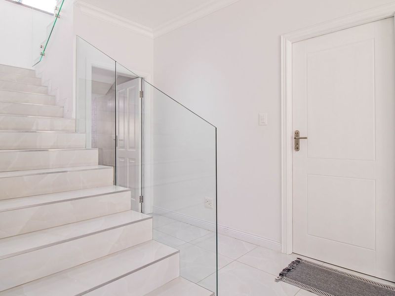 A bright, modern hallway featuring a tiled staircase with a sleek glass balustrade and clean, white finishes.