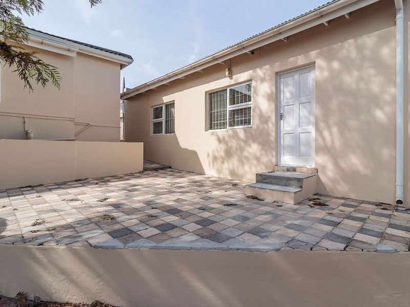 A rear exterior view featuring a paved patio area, retaining wall, and access door with security bars.
