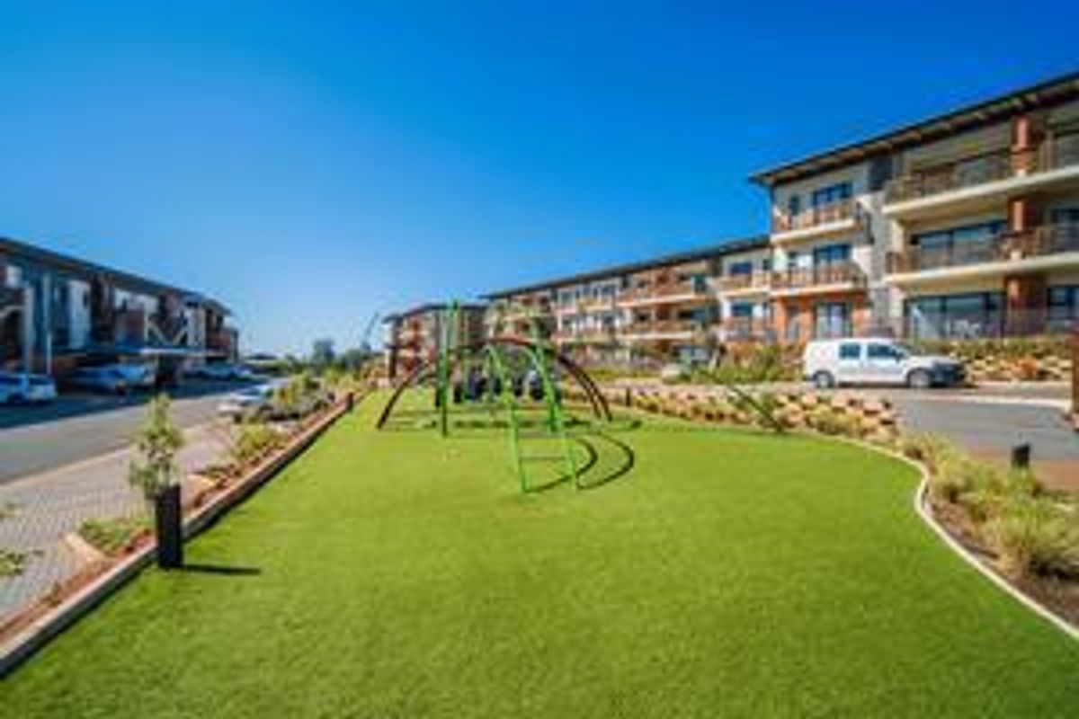 A well-maintained communal outdoor area featuring a playground, artificial lawn, and landscaped gardens situated between modern apartment blocks.