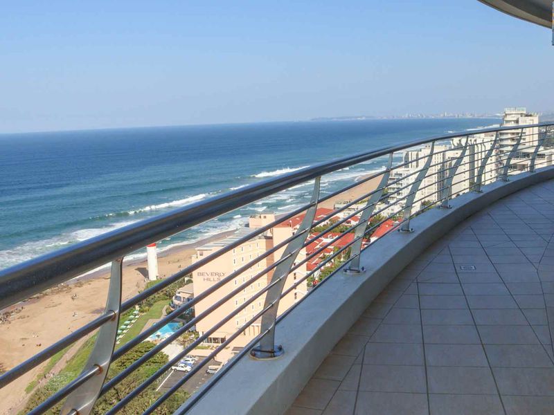 Spacious curved balcony featuring tiled flooring and a modern stainless steel railing, offering panoramic ocean and coastal views.