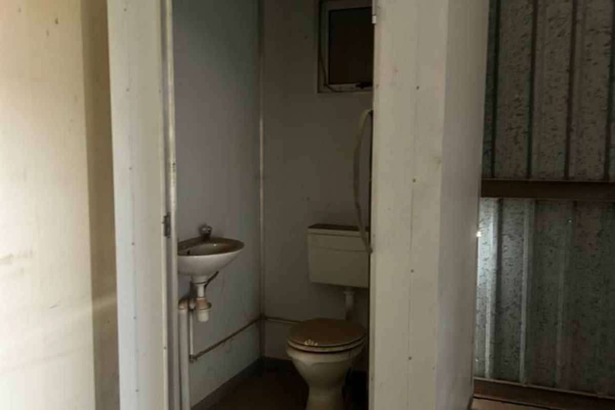 A standalone prefabricated utility unit featuring a basic toilet and small basin, situated within an industrial warehouse space.